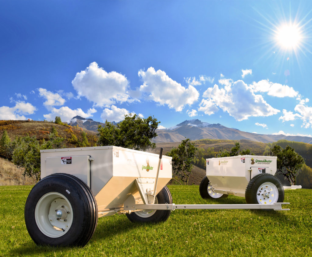 two GroundBuster brand drop spreaders sitting on grass in front of mountains